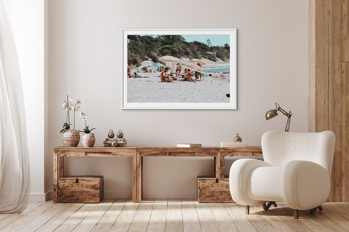 Framed coastal photograph above a timber sideboard beside a white armchair