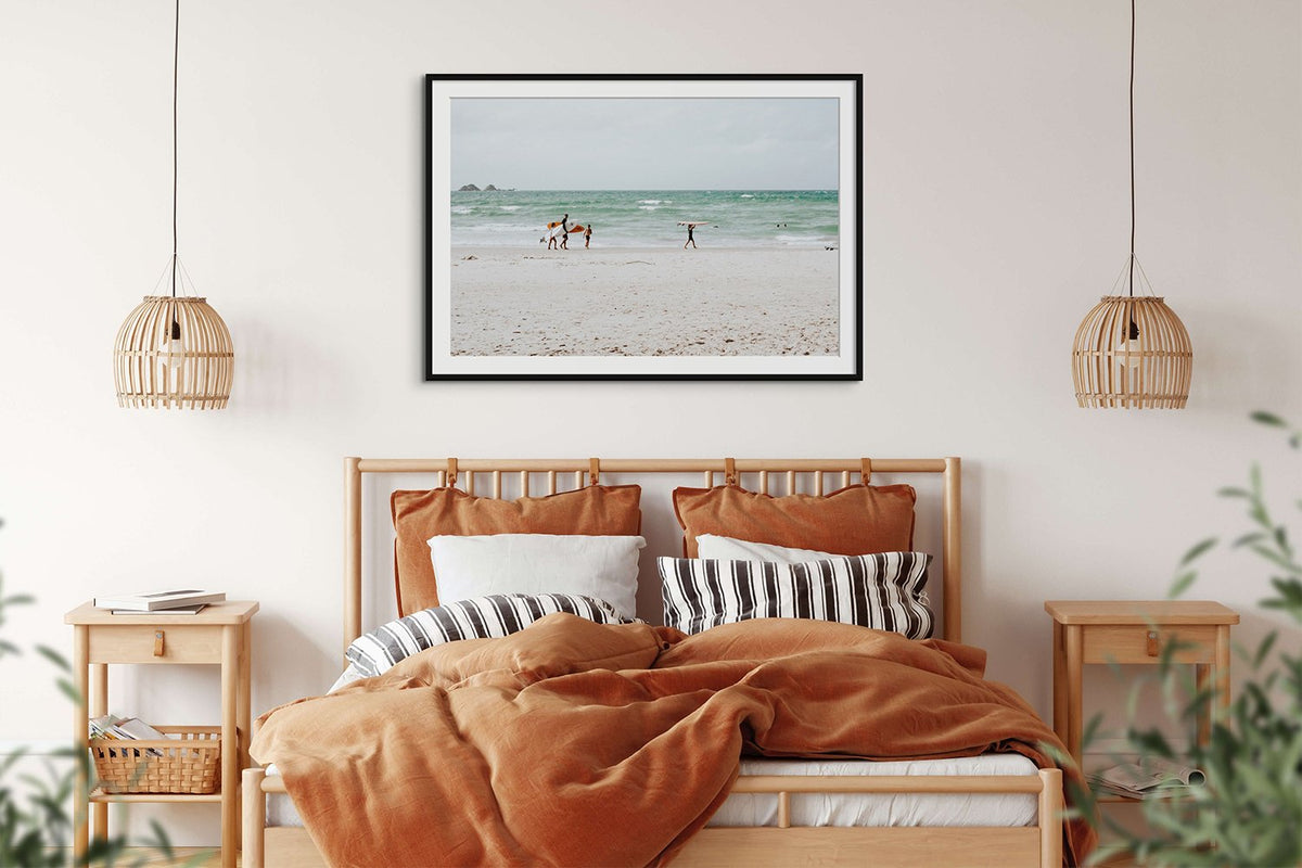 Framed coastal photograph above a bed with rust-coloured bedding and timber lamps