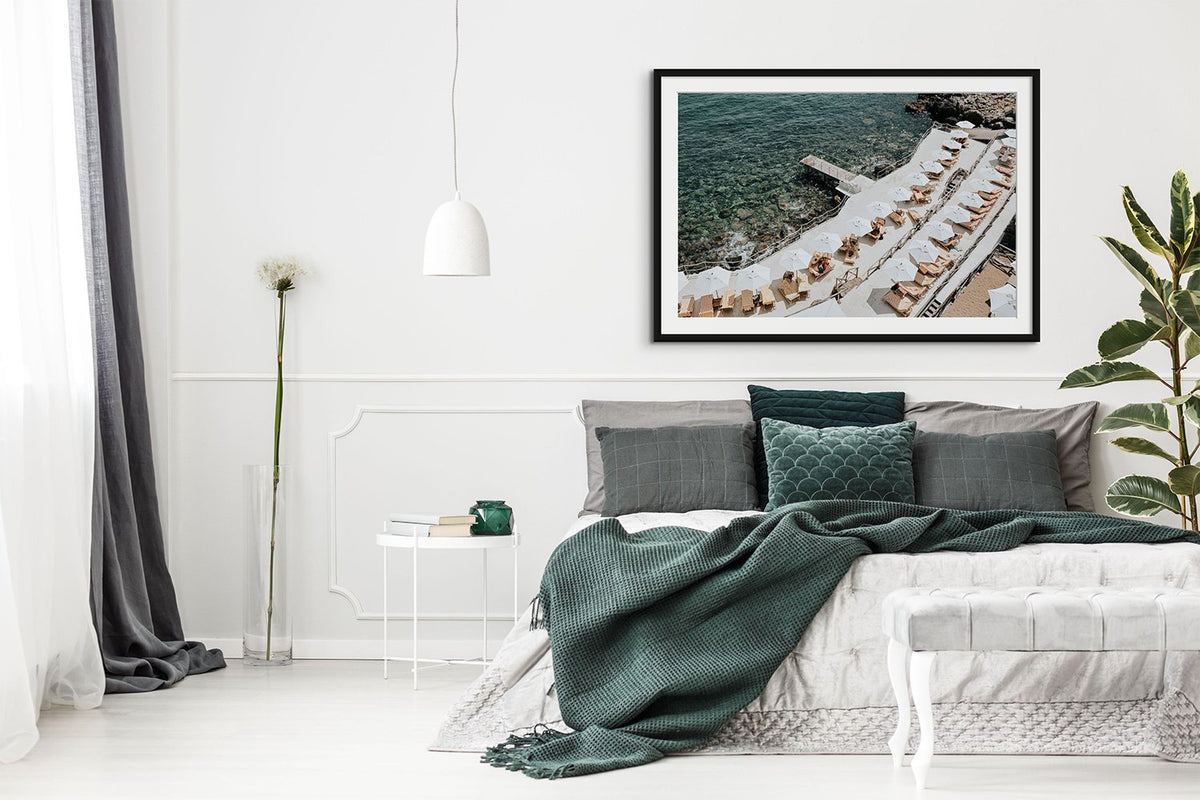 Framed coastal photograph above a bed with dark green bedding in a bright bedroom