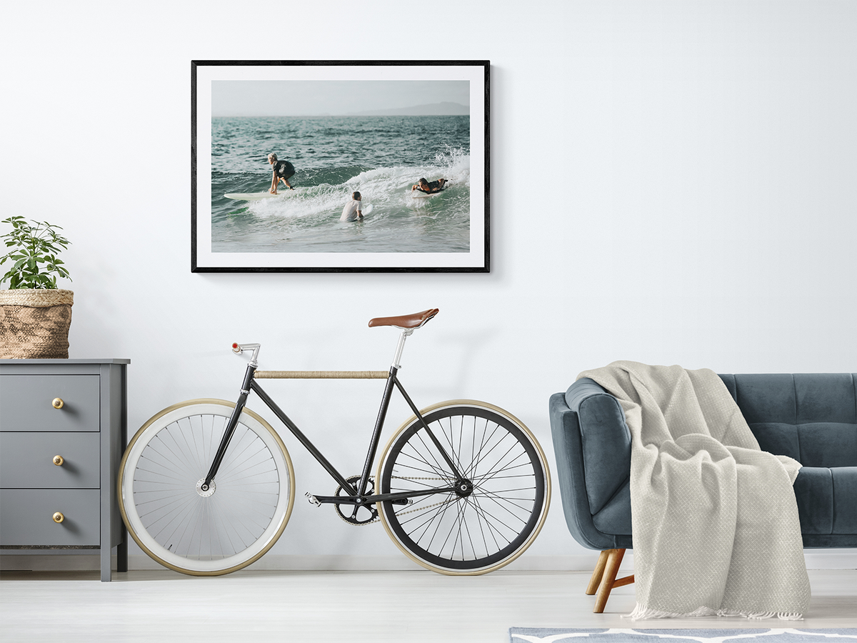 The Boys of Summer Byron Bay surf fine art print in a black frame styled in a relaxed coastal living room with a bicycle and linen throw
