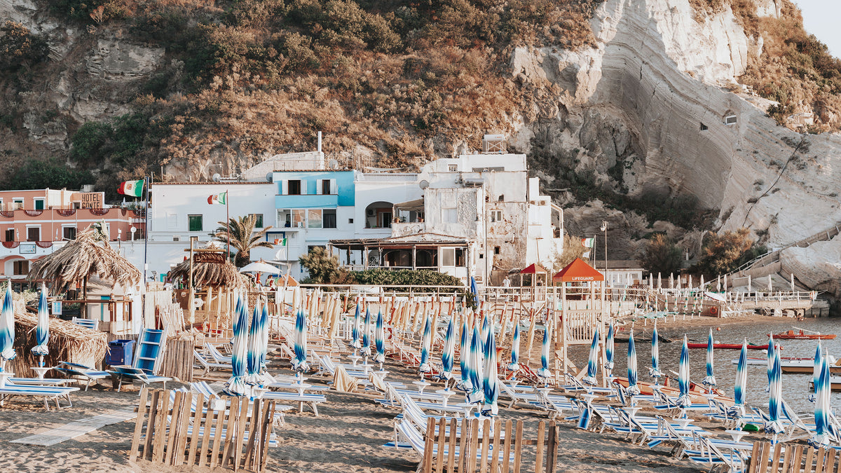 Blue loungers and umbrellas on Sant'Angelo beach in Ischia, below white houses and a pale cliff