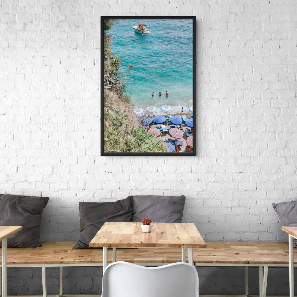 Vitamin Sea Positano, turquoise shallows, swimmers and striped umbrellas framed by greenery, framed print styled in a room